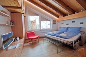 A modern bedroom with a loft bed, a red chair, a desk with a computer, and a window overlooking a building.