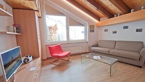 A modern living room with a wooden ceiling, a red chair, a beige sofa, a glass coffee table, and a TV on a wooden stand. - Studio Riffelsee (Zermatt)