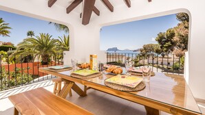 Un espace repas avec une table en verre et des bancs, offrant une vue panoramique sur la côte, avec des palmiers et un ciel dégagé.