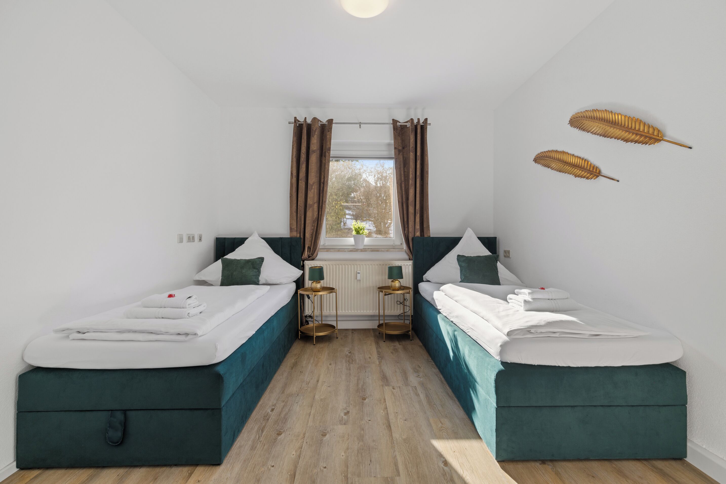 A room with two single beds, a window with curtains, a radiator, and a small table with a lamp.