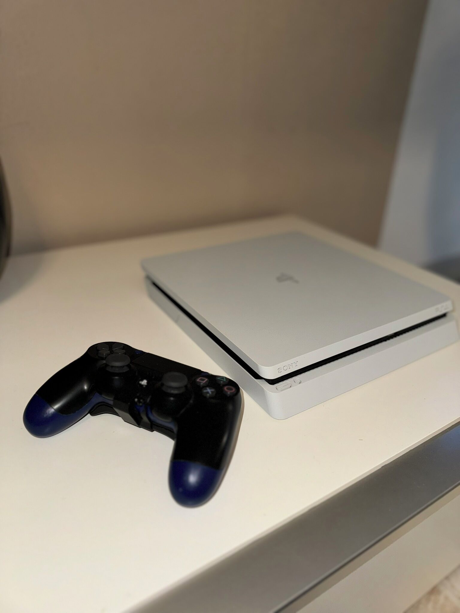 Ein weißer Nachttisch mit einem PlayStation-Controller und einer weißen Xbox Series X Konsole.