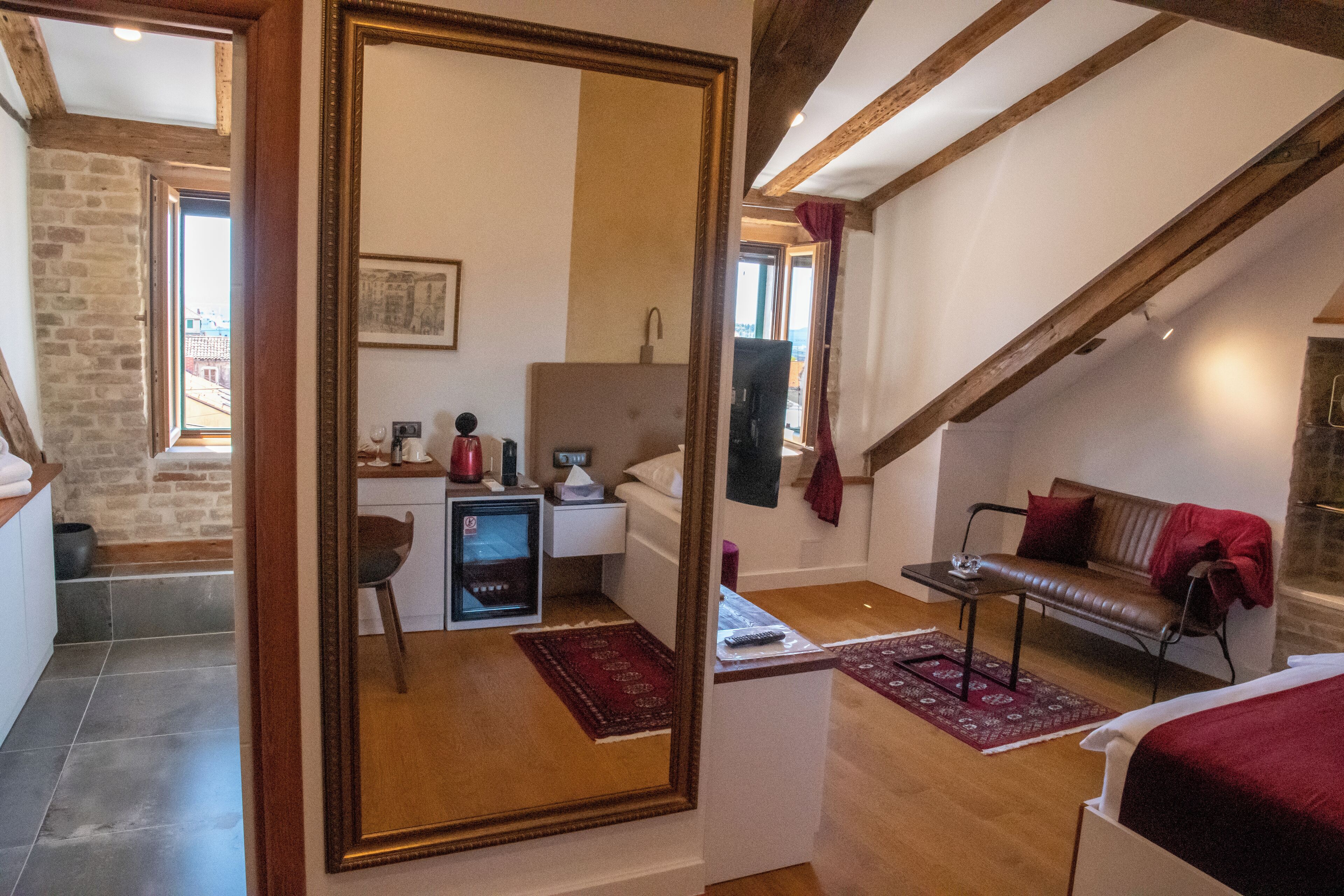 A room with a bed, a desk, a chair, a red rug, and a large mirror.