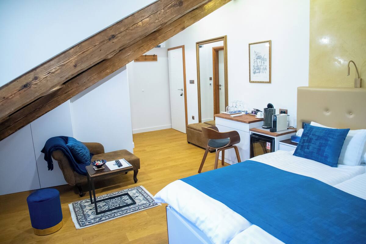 Quarto com cama grande, escrivaninha e cadeira. Apresenta um vigamento de madeira, um banquinho azul e um tapete pequeno.