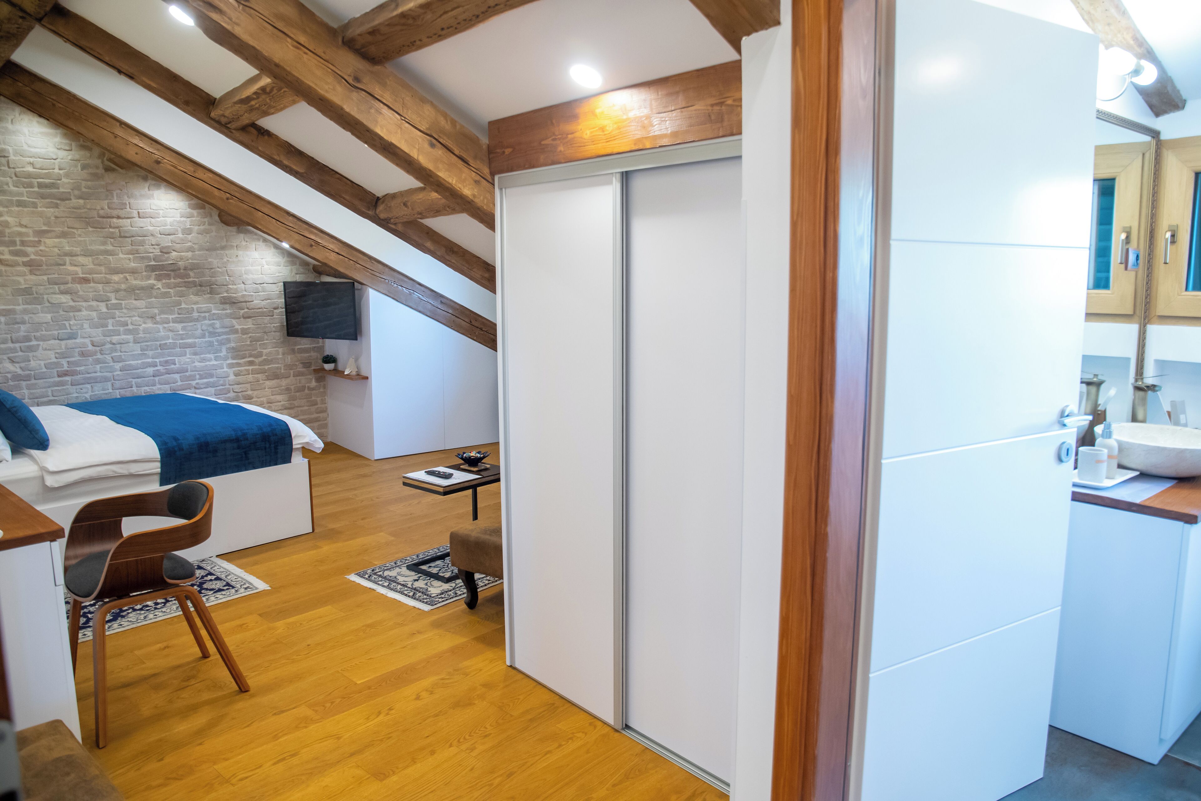 A modern bedroom with a lofted sleeping area, wooden beams, a brick wall, and a white wardrobe.