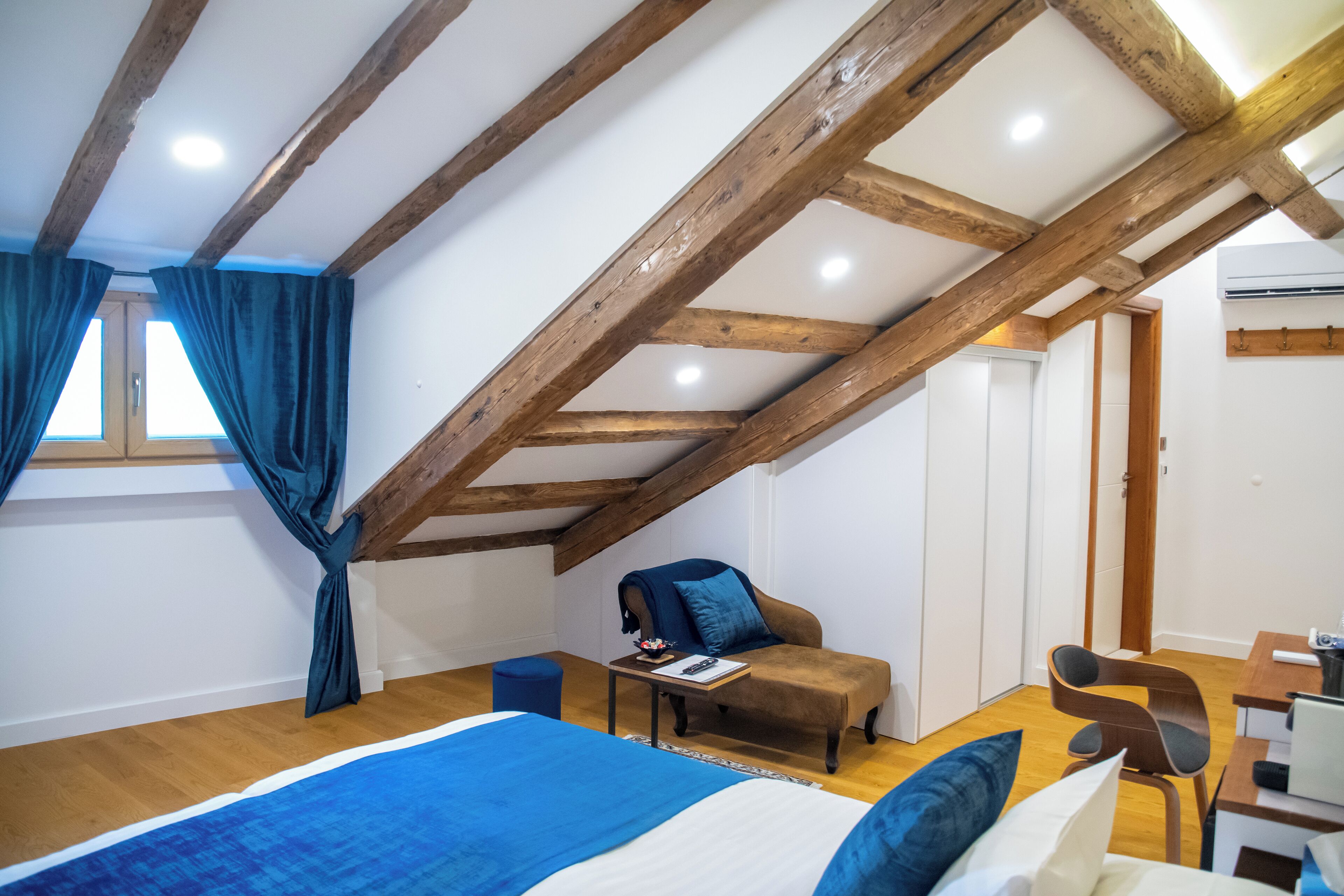 A bedroom with a sloped ceiling, wooden beams, a bed with blue bedding, a chair, a small table, and a desk with a chair.