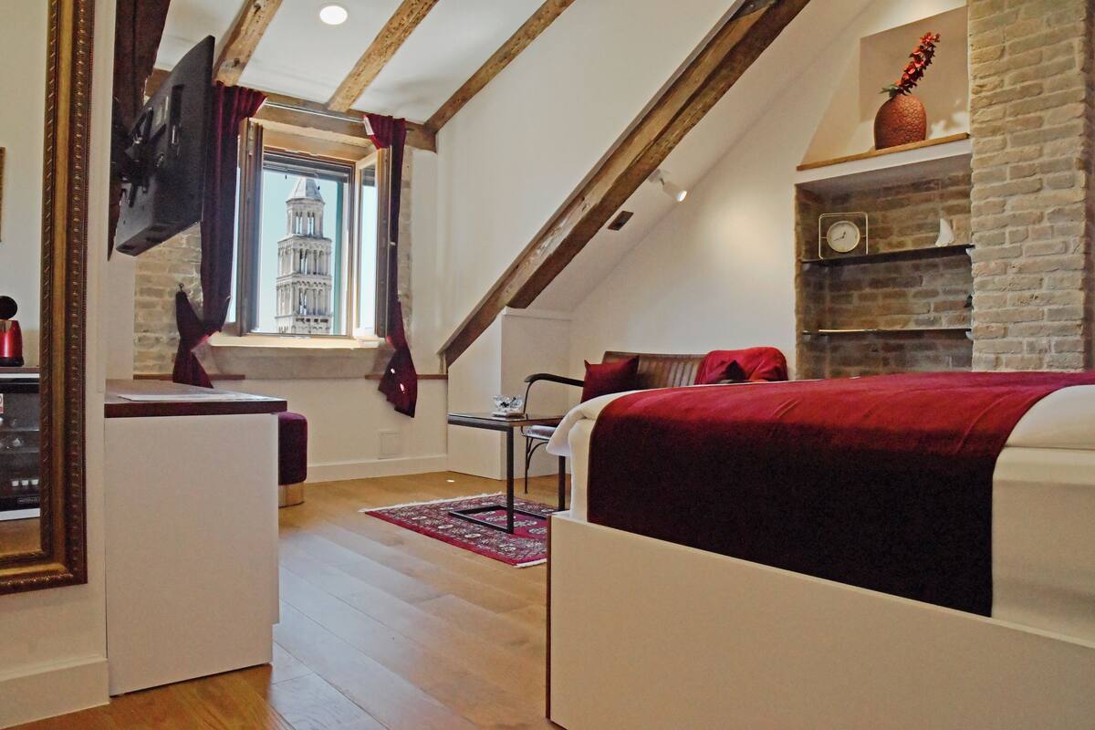 Quarto com teto inclinado, cama com manta vermelha, televisão fixada na parede, janela com vista para um edifício e lareira de tijolos.
