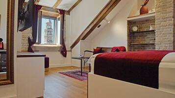 A bedroom with a sloped ceiling, a bed with a red blanket, a TV mounted on the wall, a window with a view of a building, and a brick fireplace.