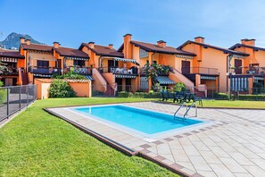 A swimming pool with a tiled deck, surrounded by a well-maintained lawn and a row of orange buildings with balconies.