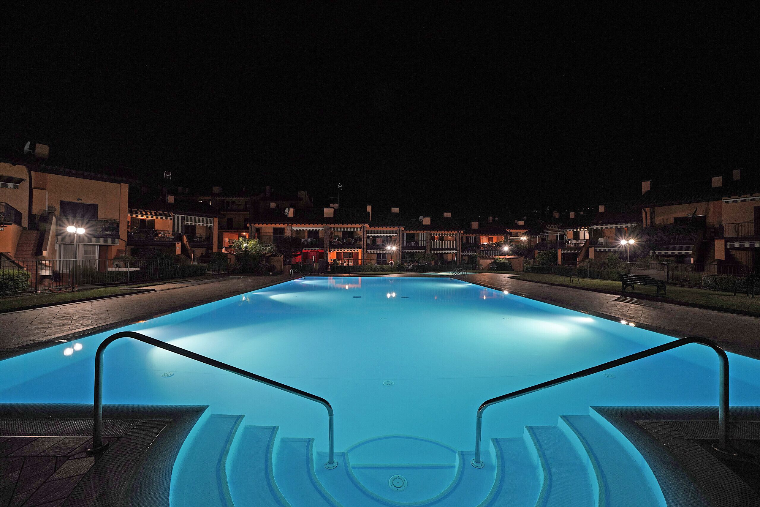 Una piscina con escalones de acceso, rodeada de edificios con ventanas iluminadas por la noche.