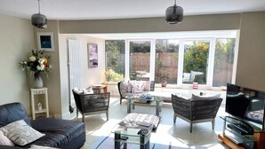 Un salon spacieux comprenant un canapé, des fauteuils, une table basse et un téléviseur. De grandes fenêtres offrent une vue sur l’extérieur.