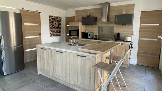 A modern kitchen with a central island, stainless steel appliances, wooden cabinetry, and a flat-screen TV mounted above the oven. - Maison Familiale Avec Jardin (Avelin)