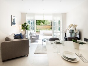 A modern living room with a grey sofa, a dining table set with plates and cutlery, and a large window offering a view of a garden. - Little Venice Charm Cozy One-bedroom by Sojo Stay (London)