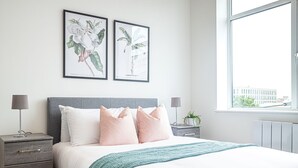 A modern bedroom with a grey headboard, white bedding, and a teal throw blanket. There are two framed botanical prints on the wall, a bedside table with a lamp, and a window with a view of buildings. - Two-bedroom Near Cliveden House by Belvilla (Slough)
