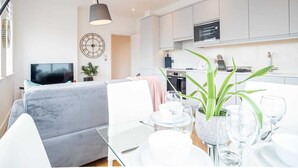 A modern living room with a grey sofa, a glass dining table set with plates and glasses, a large clock on the wall, and a kitchen area with white cabinets and appliances. - Two-bedroom Near Cliveden House by Belvilla (Slough)