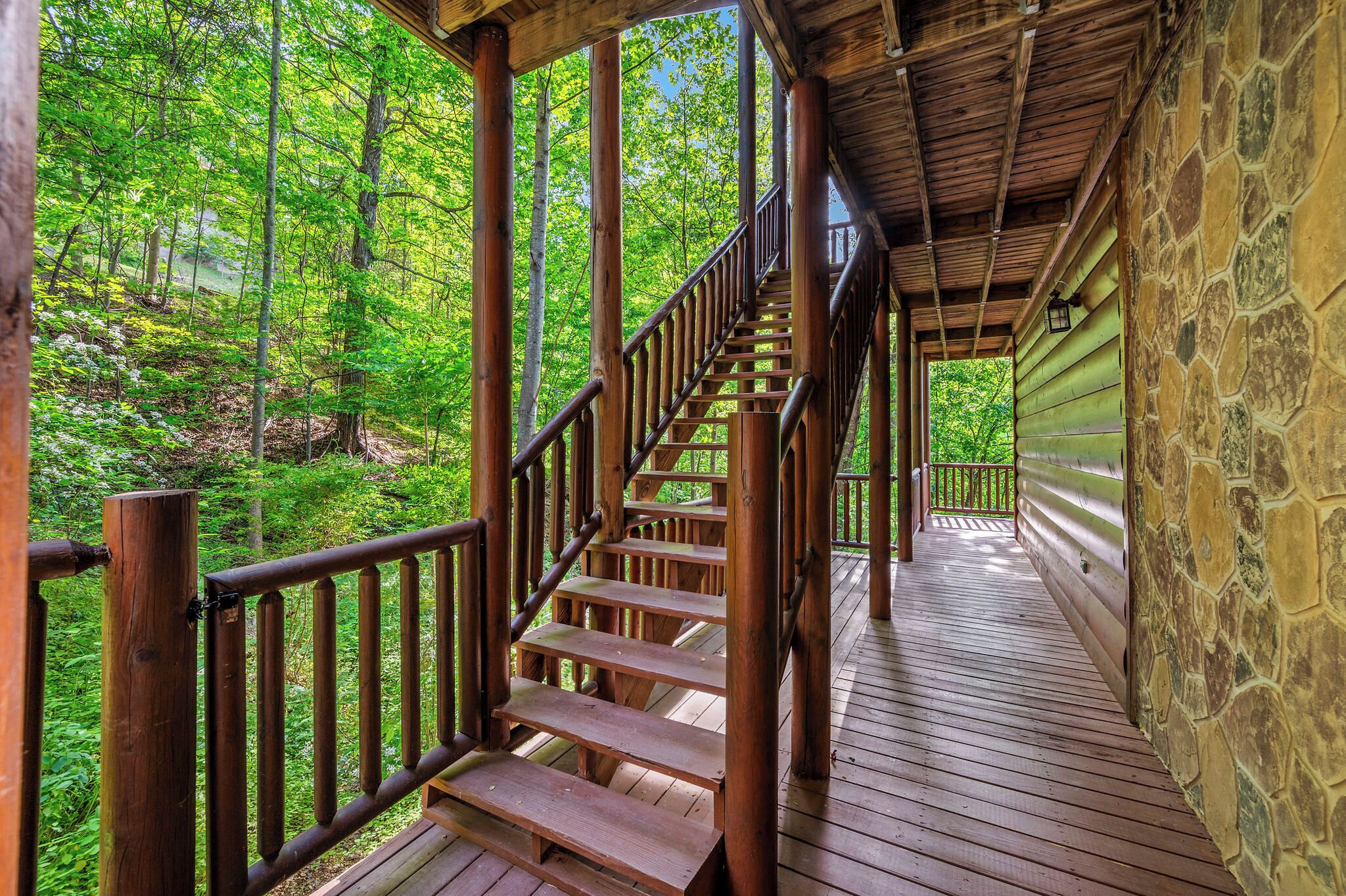 Eine Holztreppe führt zu einer überdachten Terrasse mit Steinmauern, umgeben von bewaldetem Gelände.