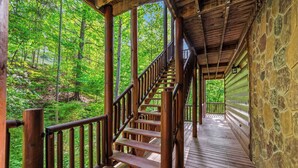 A wooden staircase leading to a covered deck with stone walls, surrounded by a forested area.