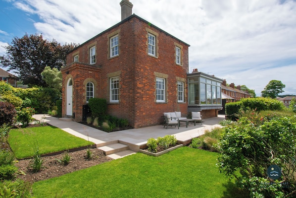 A two-story brick house with a front porch, a small garden, and a patio with outdoor furniture.