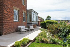 Un bâtiment en briques doté d’un patio aménagé avec du mobilier d’extérieur et d’un jardin où poussent diverses plantes et fleurs.