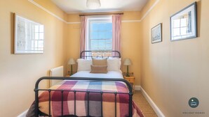 Une chambre à coucher avec un lit, une fenêtre avec des rideaux, un radiateur, deux tables de chevet avec des lampes et des tableaux encadrés accrochés aux murs.