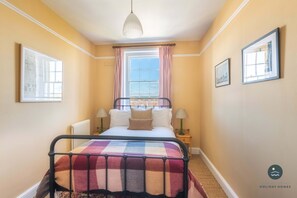 Un dormitorio con cama, ventana con cortinas, radiador, dos mesitas de noche con lámparas y cuadros en las paredes.