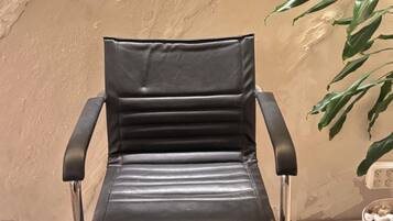 Silla de cuero negro con patas metálicas, ubicada sobre un piso moteado, frente a una pared beige con textura y una planta a la derecha.