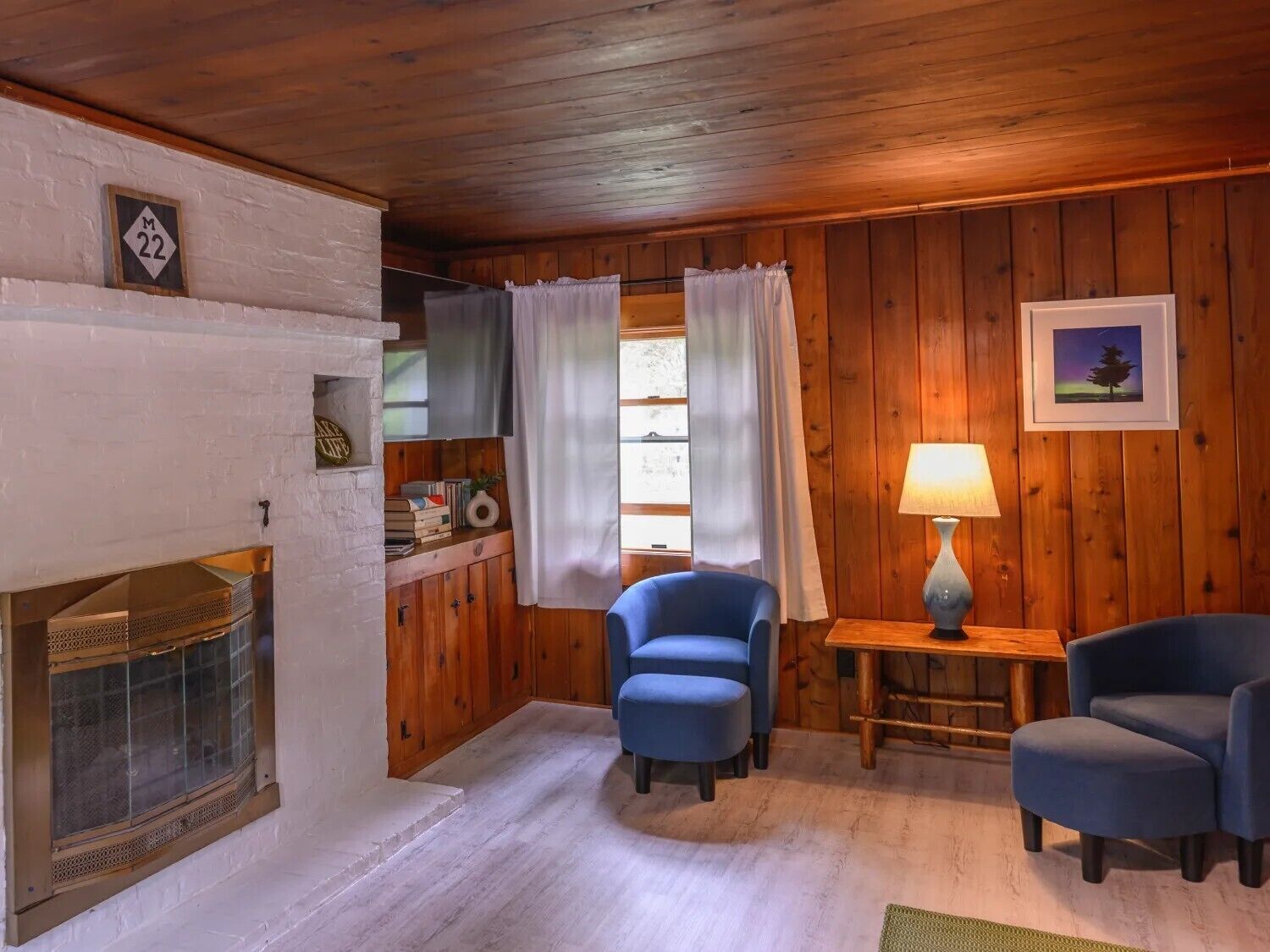 Ein gemütliches Blockhaus-Interieur mit Kamin, Holzwänden, zwei blauen Sesseln, einem kleinen Tisch mit Lampe und einem Fenster mit Vorhängen.
