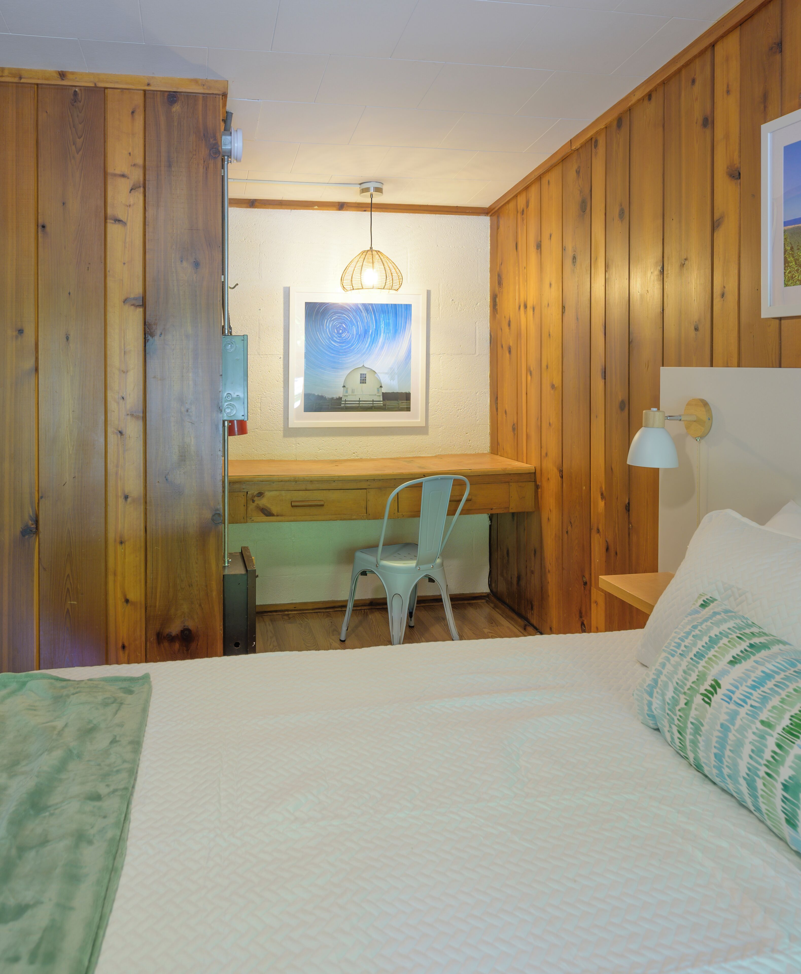 Une chambre avec un bureau en bois, une chaise, un lit recouvert d’une couverture verte, une table de chevet avec une lampe et un tableau encadré au mur.