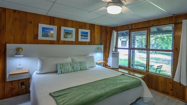 A bedroom with a large bed, wooden walls, a window with a view of the outdoors, and a ceiling fan.