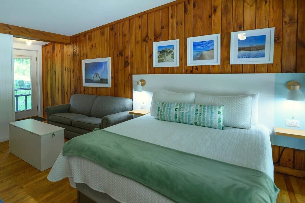 A bedroom with a wooden paneled wall, a bed with a green blanket, a grey sofa, and three framed pictures.
