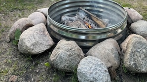 A metal fire pit with rocks around it, two Adirondack chairs, and a wooden structure in the background.