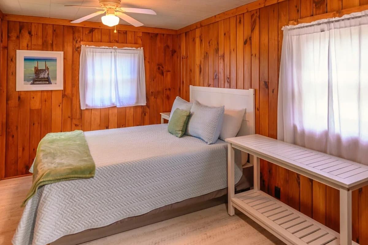 A wooden-paneled room with a bed, a white headboard, a bedside table, and a ceiling fan.