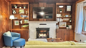 A cozy living room with a fireplace, a television, wooden shelves with books and decorations, and a blue armchair.