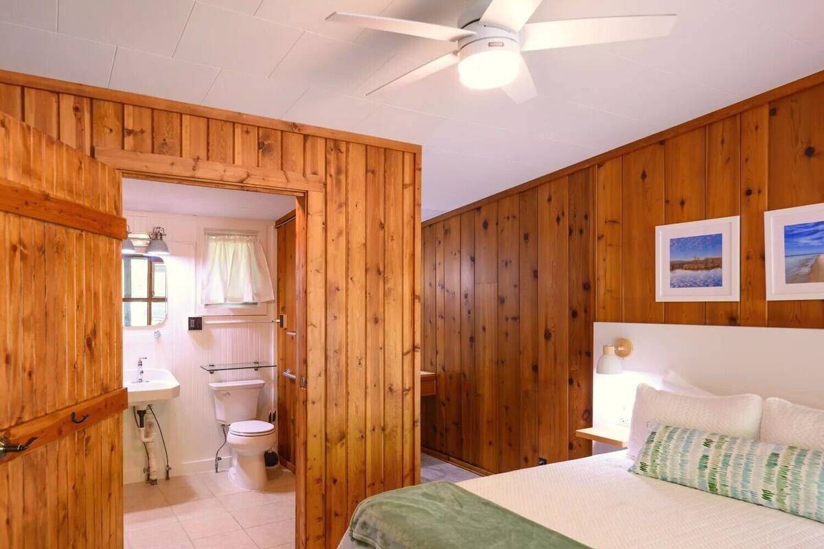 A wooden-paneled room with a bed, a ceiling fan, and a bathroom visible through an open door.