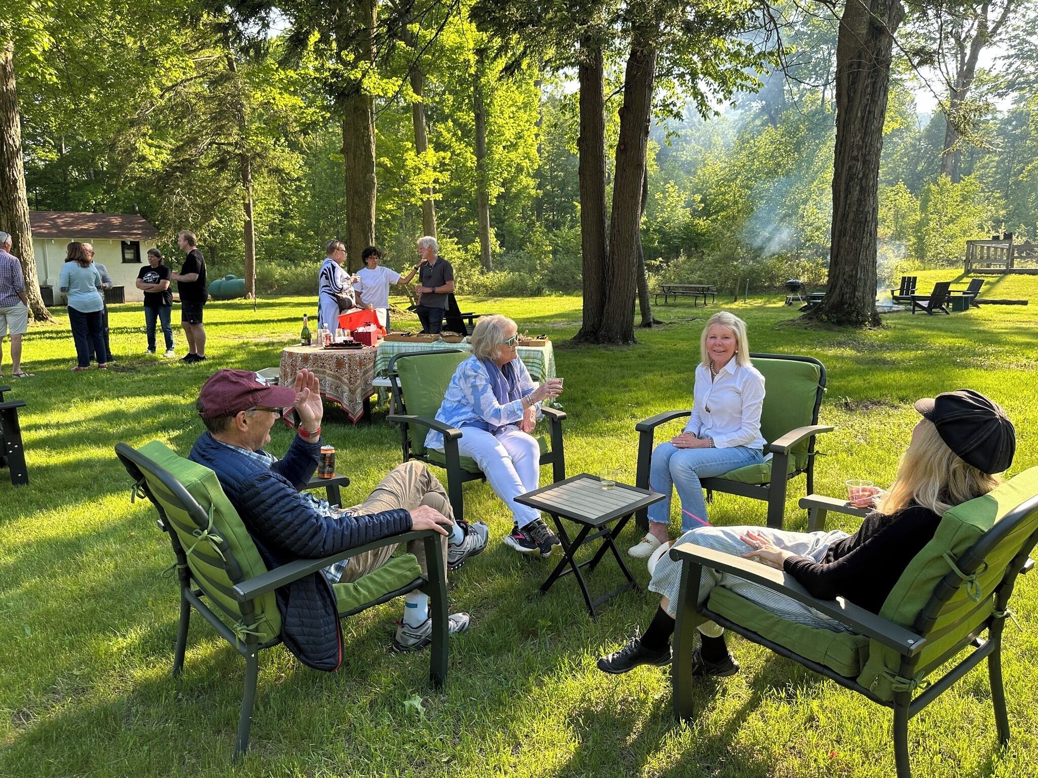 A group of people seated on green chairs in a grassy outdoor area with trees in the background.