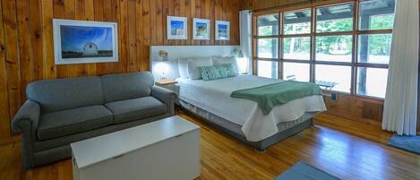 Una habitación con un revestimiento de madera, una cama con cabecera blanca, un sofá gris, una mesita de noche blanca y un ventanal con cortinas.
