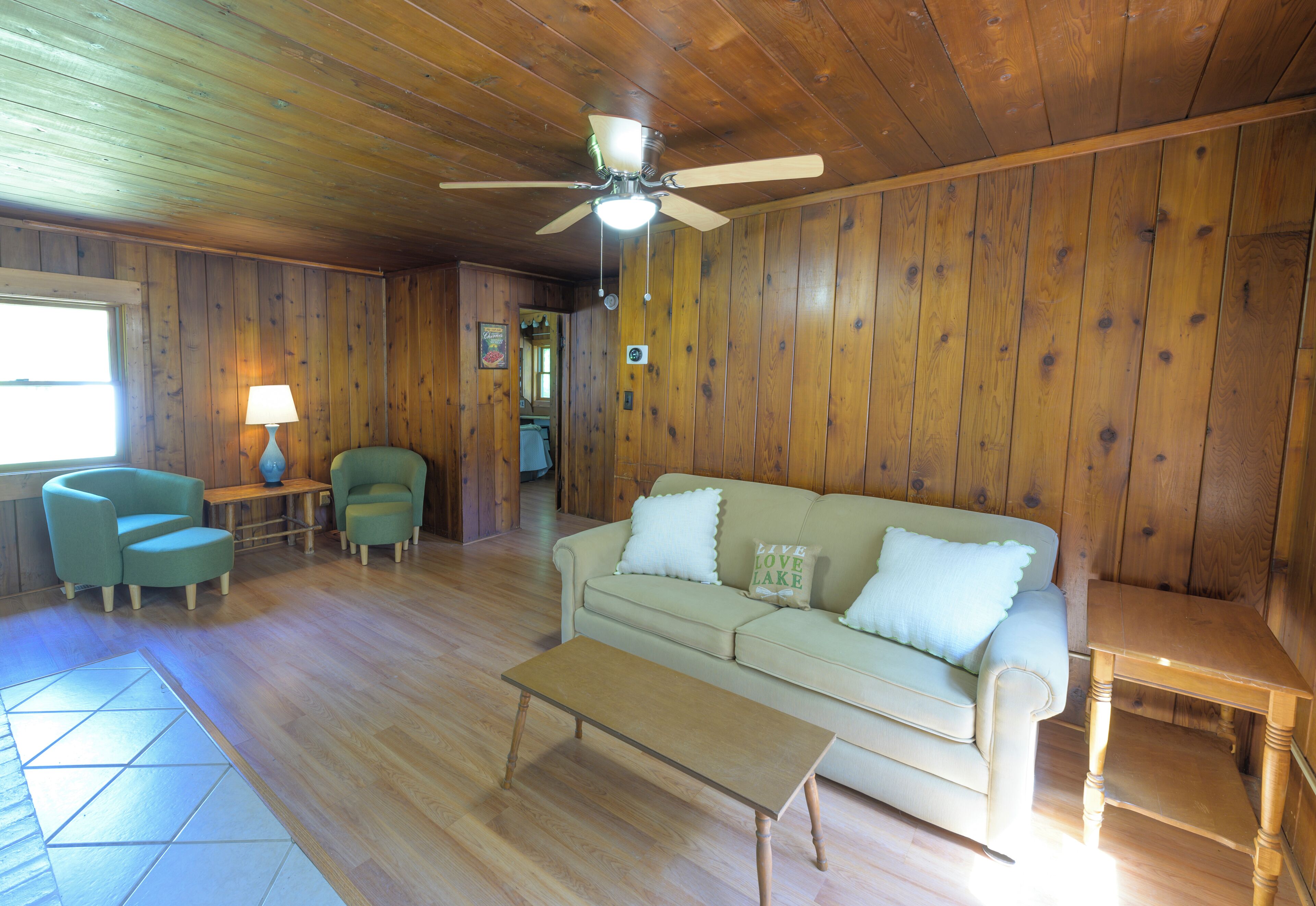 Una habitación con paneles de madera, un sofá color beige, dos sillones, una mesa de centro y un ventilador de techo.