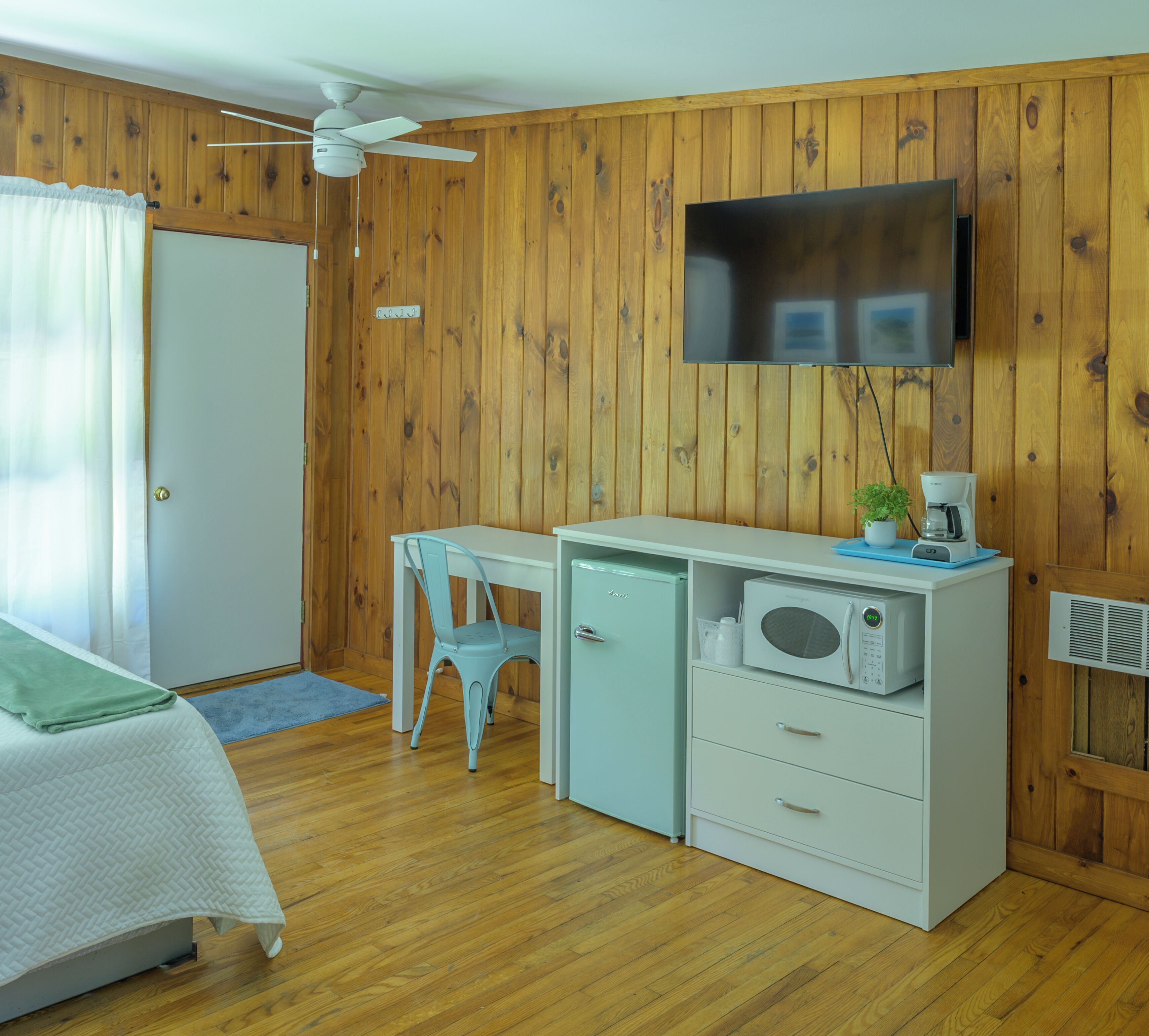 A room with wooden walls, a bed with a white bedspread, a desk with a chair, a microwave, and a television mounted on the wall.