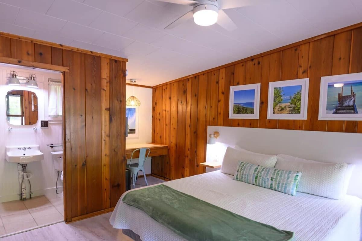 A bedroom with wooden paneling, a bed with white bedding, a desk with a chair, and a bathroom with a sink and mirror.