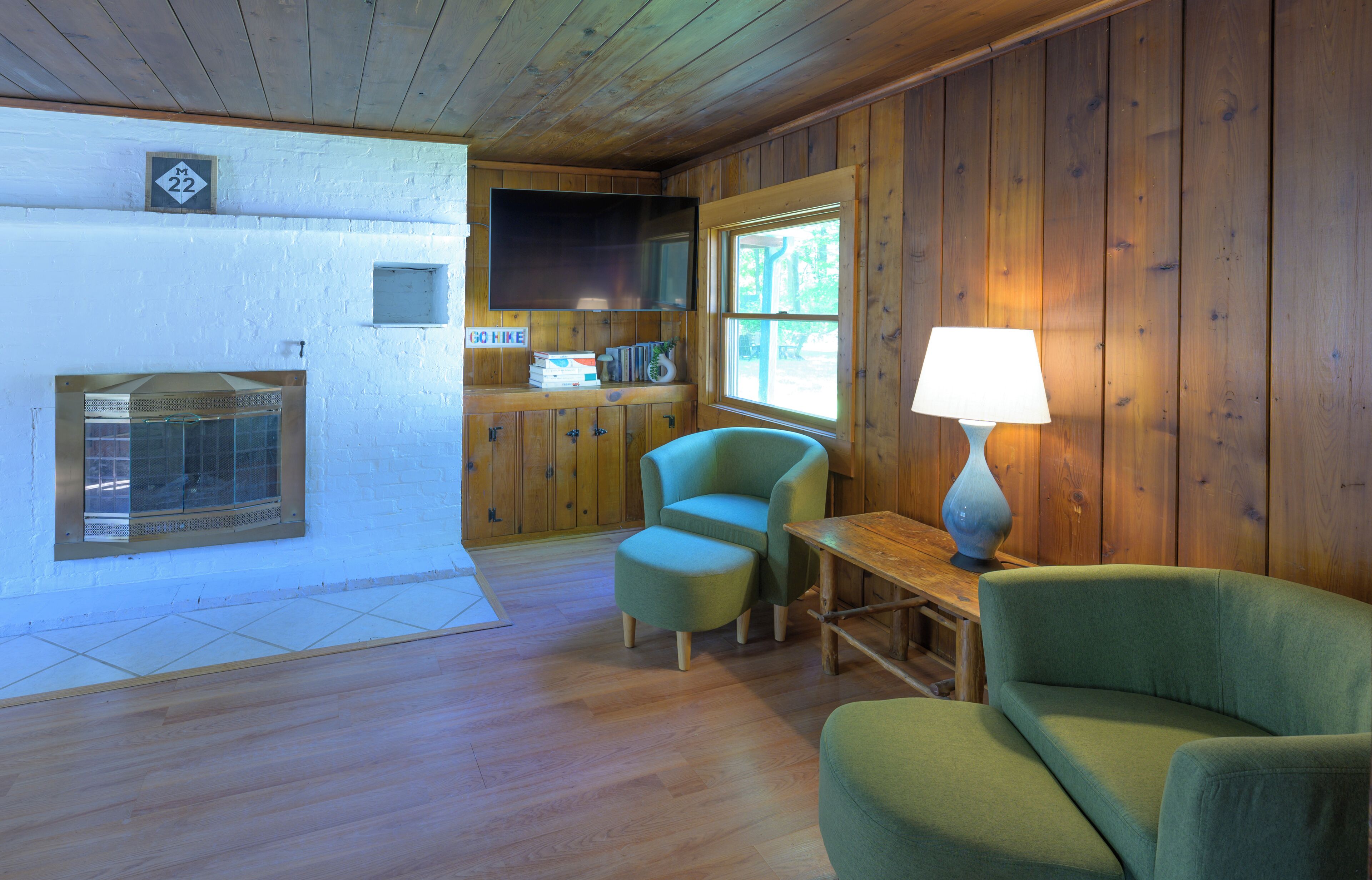 Interior acogedor de una cabaña con paredes y suelos de madera, un televisor sobre la chimenea y dos sillones verdes.
