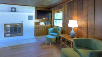 A cozy cabin interior with wooden walls and floors, a television mounted above a fireplace, and two green armchairs.