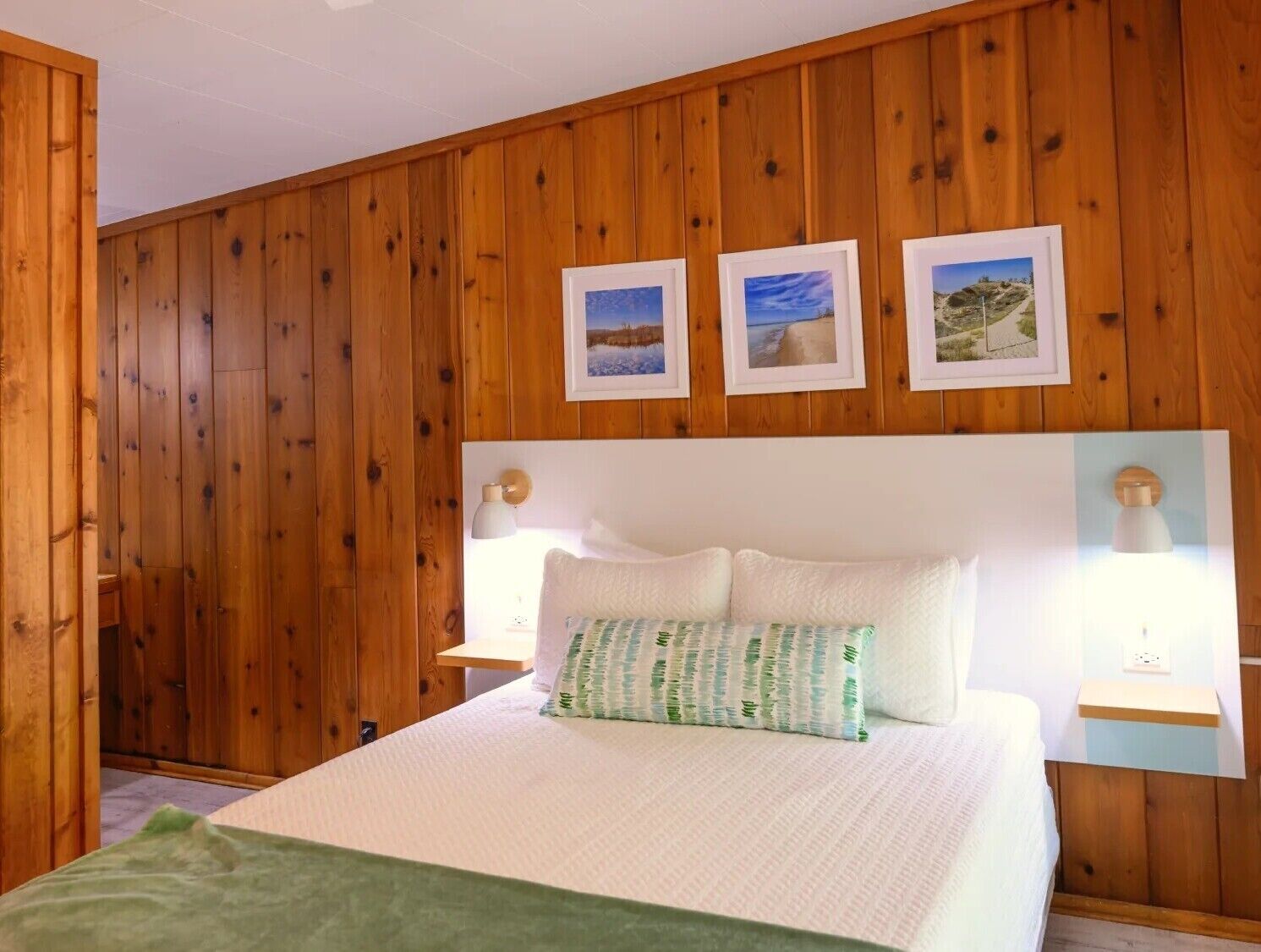 Une chambre avec un mur lambrissé en bois, un lit avec une literie blanche, deux lampes de chevet et trois tableaux encadrés au-dessus du lit.