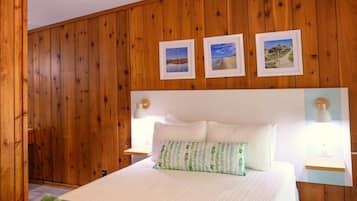 Une chambre avec un mur lambrissé en bois, un lit avec une literie blanche, deux lampes de chevet et trois tableaux encadrés au-dessus du lit.