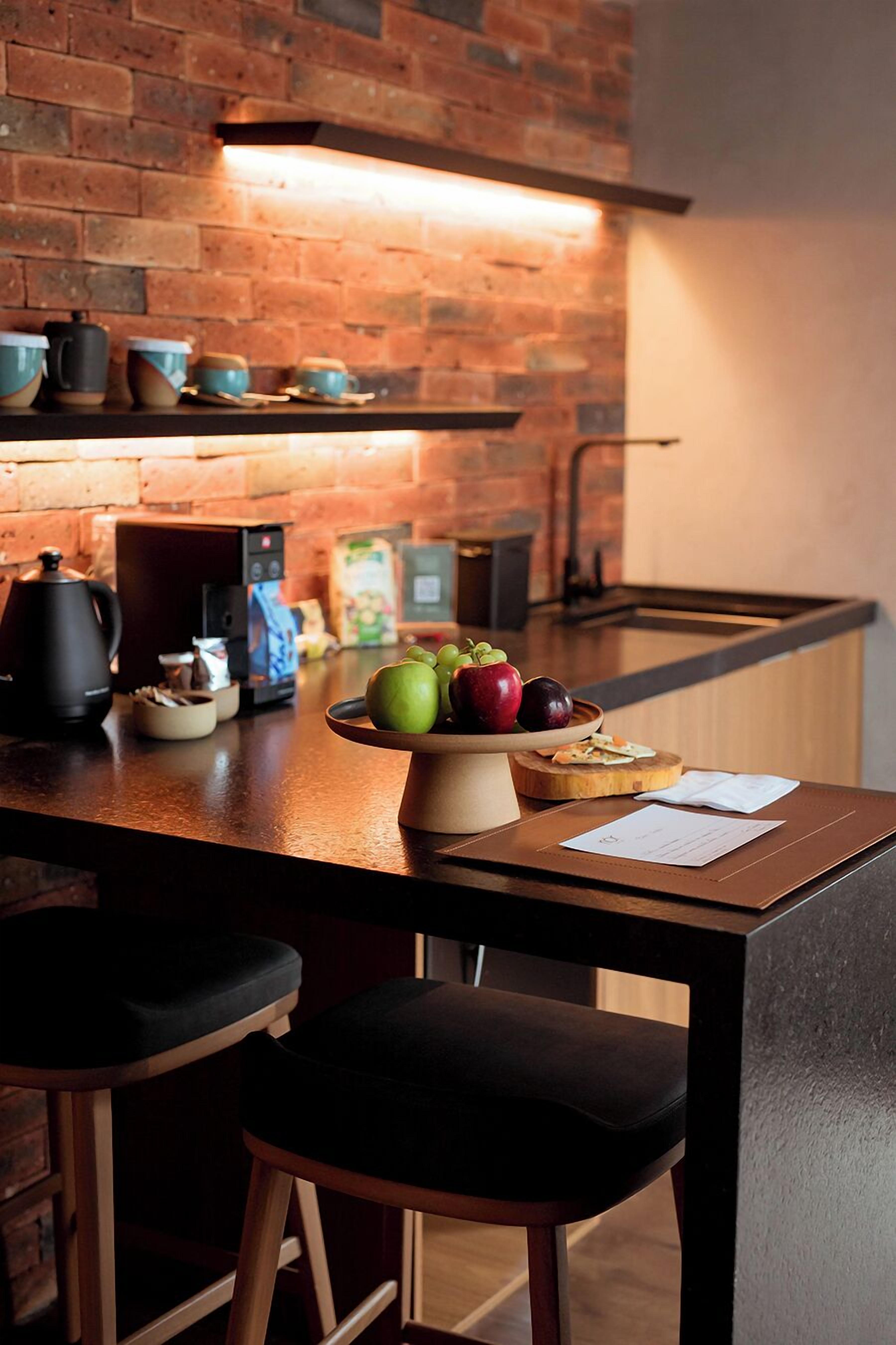 A kitchen with a brick wall, a coffee machine, a bowl of fruit, and bar stools.