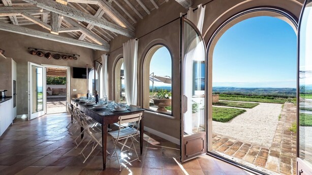 A spacious dining area with a long table set for a meal, large arched windows, and a view of a landscaped garden.