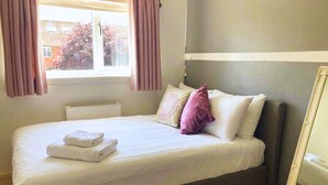 A single bed with white linens and a folded towel, a window with curtains, and a mirror on the wall. - Chichester House I Secure Garden I Parking I Close to Goodwood (Chichester)