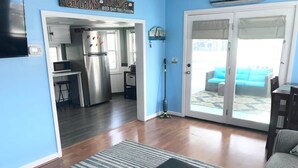 A living room with a black ottoman, a striped rug, and a view of a kitchen and dining area through a glass door.
