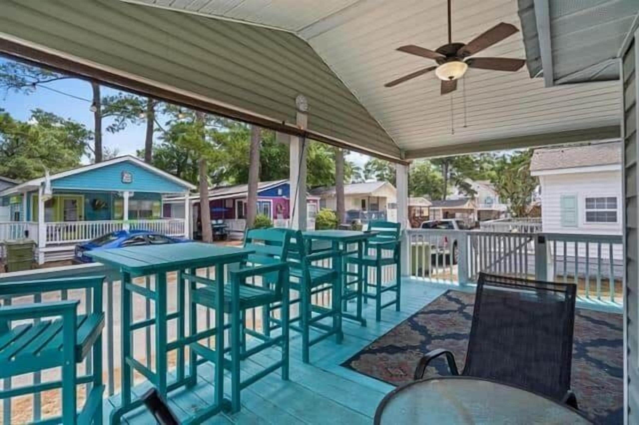 A covered outdoor seating area with tables and chairs, a ceiling fan, and a view of neighboring houses.