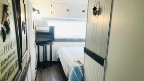 A narrow corridor with a bed on the right, a small table with a TV, and a sign on the left.