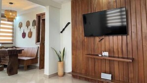 A modern living room with a wooden wall panel, a flat-screen TV, a dining area with a wooden table and chairs, and a potted plant. - Monie's Quarters (Kumasi)
