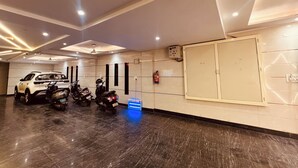 Un espace de stationnement où se trouvent un VUS blanc et plusieurs scooters, ainsi qu’un extincteur et un panneau de commande fixé au mur.